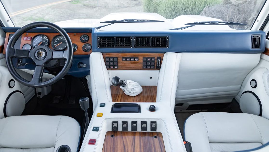vehicle interior with steering wheel, analog gauges, wood trim dashboard, manual gear shifter, white seats.