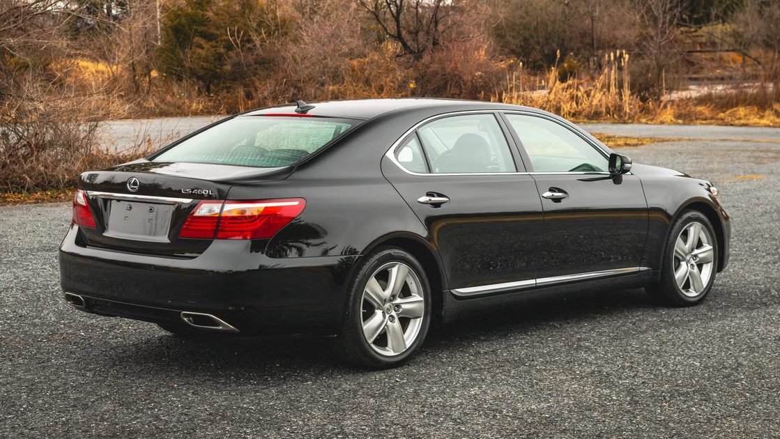 rear three quarter view black sedan parked on pavement with trees and brush in background.