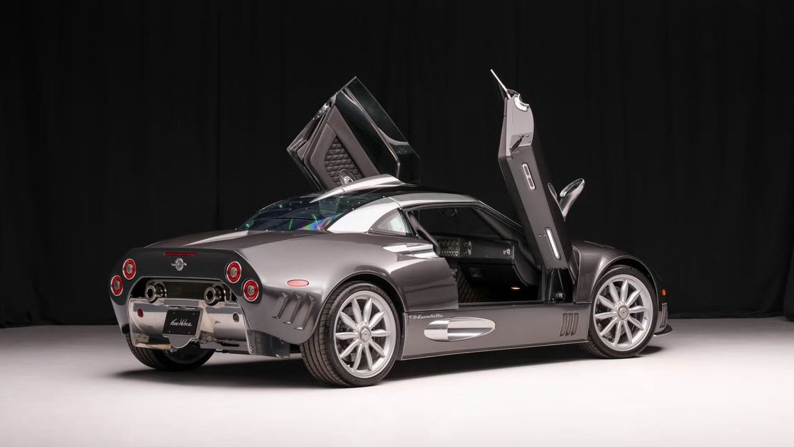 rear three quarter view silver sports coupe with upward opening doors in studio with black curtain backdrop.