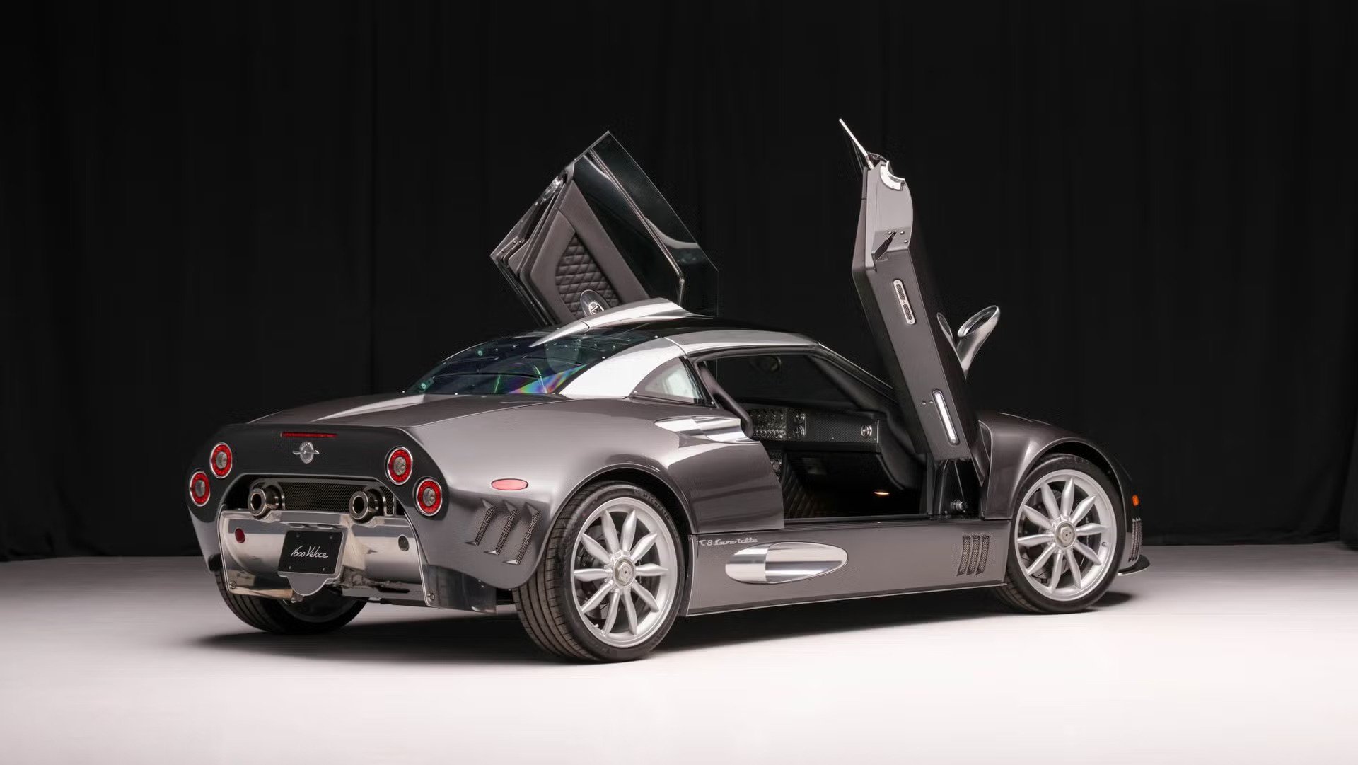 rear three quarter view silver sports coupe with upward opening doors in studio with black curtain backdrop.